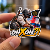 A close-up of a hand holding a die-cut sticker featuring a PUBG-style character with helmets and a rifle, plus bold text reading "ONXON?!" and a small "Winner Winner Chicken Dinner" badge. The background is softly blurred.