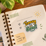 A spiral-bound notebook with a decorative sticker that reads 'Tenang is Good!' in yellow, green, and white. The notebook has a dotted grid cover, decorated with green washi tape, a yellow sticky note that says 'Take a deep breath', and is styled on a wooden surface with a plant leaf in the background. FOR ILLUSTRATION PURPOSES ONLY.