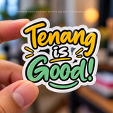 A hand holds a die-cut sticker that reads “Tenang is Good!” in bold yellow and green bubble lettering with black outlines and a white border, against a blurred indoor background.